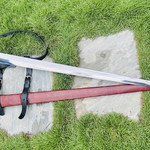 Op de afbeelding: Een lang, zilveren zwaard met een zwarte handgreep en pareerstang, rustend op een rode leren schede. Het zwaard is vastgezet met zwarte leren riemen en een riem. De items liggen op een stenen oppervlak met groen gras.