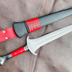 May include: A silver dagger with a red and black leather sheath. The dagger has a pointed tip and a straight blade. The sheath has a decorative design and a strap.