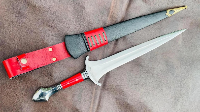 May include: A silver dagger with a red and black leather sheath. The dagger has a pointed tip and a straight blade. The sheath has a decorative design and a strap.