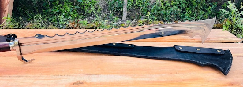 May include: A silver-colored sword with a serrated blade and a black leather scabbard. The sword has a wooden handle with a red tip.