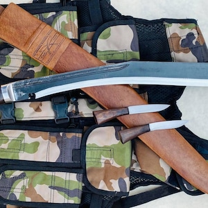 May include: A black and brown curved blade with a wooden handle and a camouflage vest. The blade is attached to the vest with straps. There are two smaller knives with wooden handles on the vest.