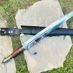 May include: A double-edged steel sword with a pointed tip and a metallic sheen. The hilt features a brown wooden grip with cross-hatching, a silver crossguard, and a pommel. It is accompanied by a black leather scabbard with a long strap and metal tip.