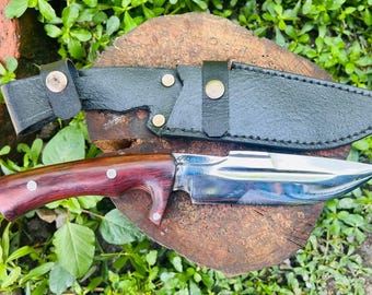 May include: A close-up of a knife with a wooden handle and a silver blade. The knife is resting on a wooden surface with a black leather sheath beside it.