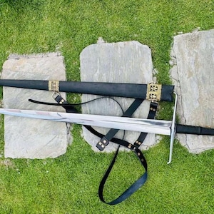 Puede incluir: Una espada plateada con una vaina de cuero negro y una empuñadura de metal dorado. La espada está sobre una superficie de hierba verde con tres piedras grises.