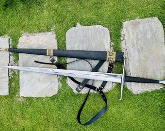 May include: A silver sword with a black leather scabbard and a gold metal hilt. The sword is lying on a green grassy surface with three gray stones.