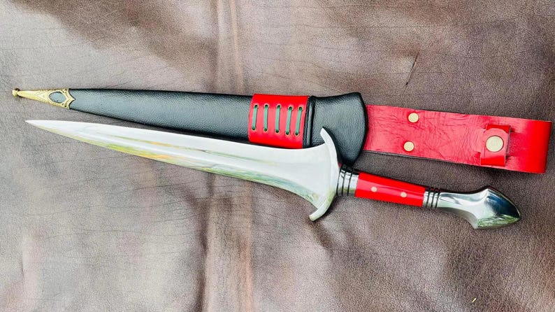 May include: A silver dagger with a red and white handle and a black leather sheath with red stitching.