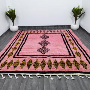 Op de afbeelding: Een roze en zwart Marokkaans tapijt met een geometrisch patroon. Het tapijt heeft een franje rand en is gemaakt van wol.