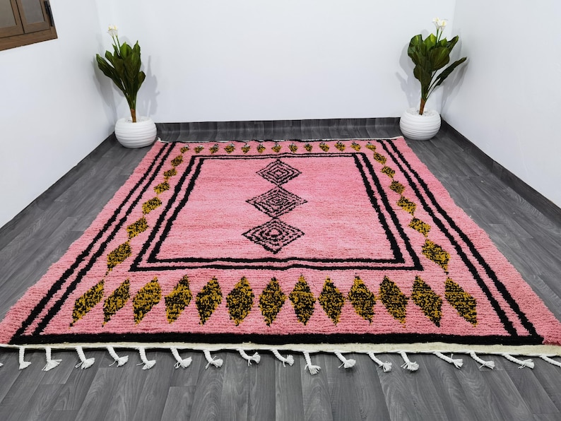 Op de afbeelding: Een roze en zwart Marokkaans tapijt met een geometrisch patroon. Het tapijt heeft een franje rand en is gemaakt van wol.