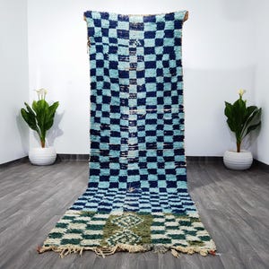 May include: A long, rectangular rug with a checkered pattern in shades of blue and white. The rug has a textured appearance and a fringed edge. The rug is displayed against a white wall with two potted plants on either side.