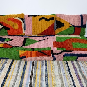 May include: A vibrant rug and cushions set with a geometric design. The rug and cushions display a mix of pink, green, yellow, orange, and black shapes. The cushions are rectangular and complement the larger rug, creating a unified aesthetic.