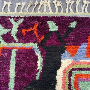 May include: A colorful rug with abstract geometric patterns in shades of purple, orange, green, blue, and white. The rug has a fringe border.