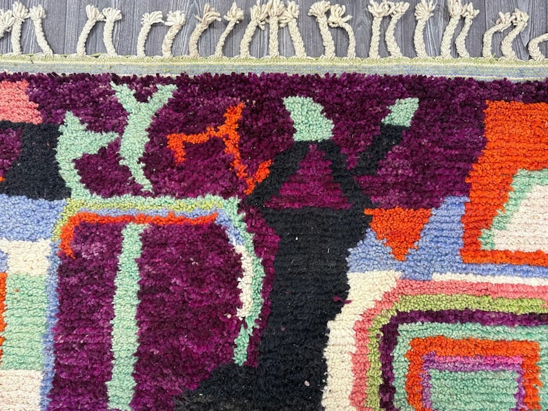 May include: A colorful rug with abstract geometric patterns in shades of purple, orange, green, blue, and white. The rug has a fringe border.