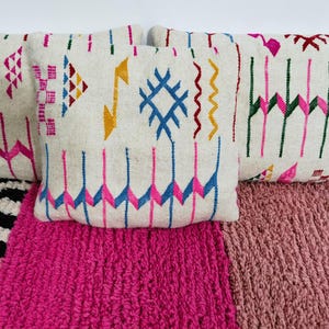 Puede incluir: Una colecci&oacute;n de cojines decorativos con coloridos patrones geom&eacute;tricos. Los cojines presentan un fondo color crema con dise&ntilde;os bordados en rosa, azul, amarillo y verde. Descansan sobre alfombras texturizadas rosas y a rayas blancas y negras.