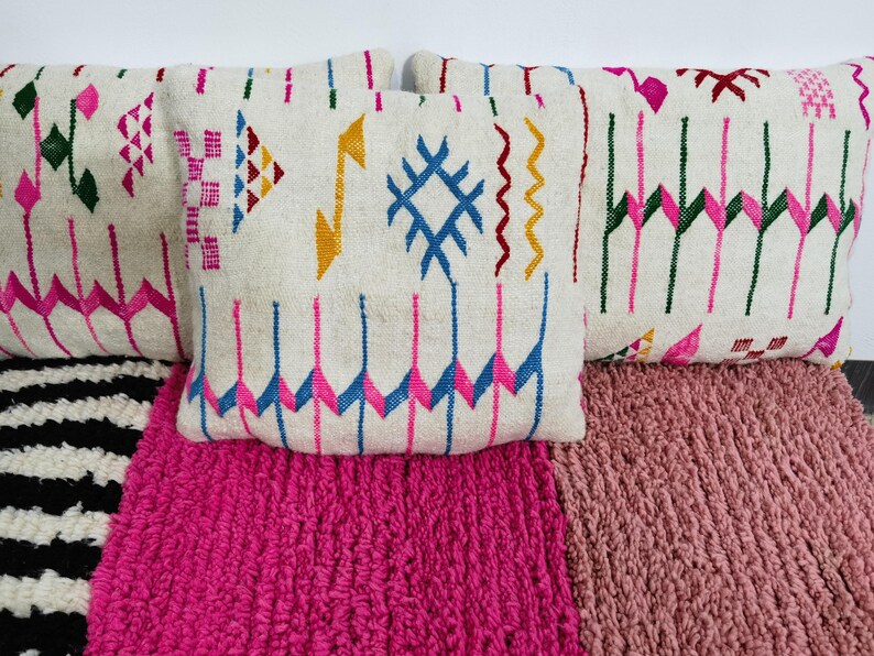 Puede incluir: Una colecci&oacute;n de cojines decorativos con coloridos patrones geom&eacute;tricos. Los cojines presentan un fondo color crema con dise&ntilde;os bordados en rosa, azul, amarillo y verde. Descansan sobre alfombras texturizadas rosas y a rayas blancas y negras.
