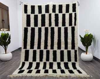 May include: A large, rectangular rug featuring a geometric design of black vertical stripes and black squares on a cream backdrop. The rug has a fringed edge and is displayed against a white wall.