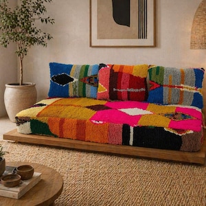 Könnte beinhalten: Ein farbenfrohes Sofa im Patchwork-Stil mit Holzsockel. Das Sofa zeigt eine lebendige Mischung aus Farben wie Gelb, Orange, Pink, Blau und Grün. Dekorative Kissen mit geometrischen Mustern liegen an der Rückseite des Sofas. Der Raum hat eine natürliche, böhmische Ästhetik.
