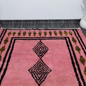 Op de afbeelding: Een roze Marokkaanse tapijt met een geometrisch patroon in zwart en goud. Het tapijt heeft een diamantvorm in het midden en een rand met een herhalend patroon.