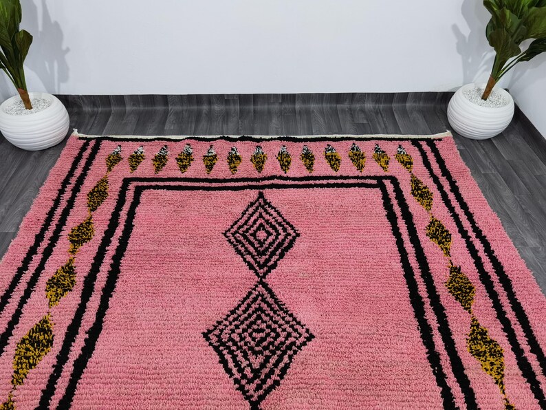 Op de afbeelding: Een roze Marokkaanse tapijt met een geometrisch patroon in zwart en goud. Het tapijt heeft een diamantvorm in het midden en een rand met een herhalend patroon.