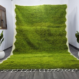 May include: A green shag rug with a white border and fringe. The rug is laid out on a gray floor.