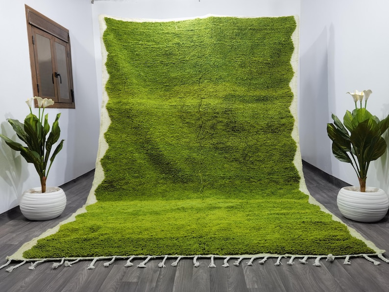 May include: A green shag rug with a white border and fringe. The rug is laid out on a gray floor.