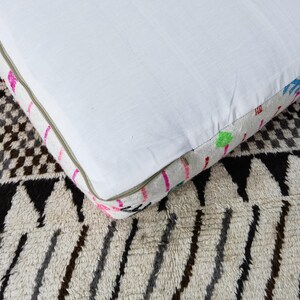 Puede incluir: Un coj&iacute;n cuadrado blanco con un borde estampado colorido. El borde presenta detalles en rosa, amarillo, verde y azul. El coj&iacute;n est&aacute; colocado sobre una alfombra estampada con dise&ntilde;os geom&eacute;tricos en negro, blanco y gris.