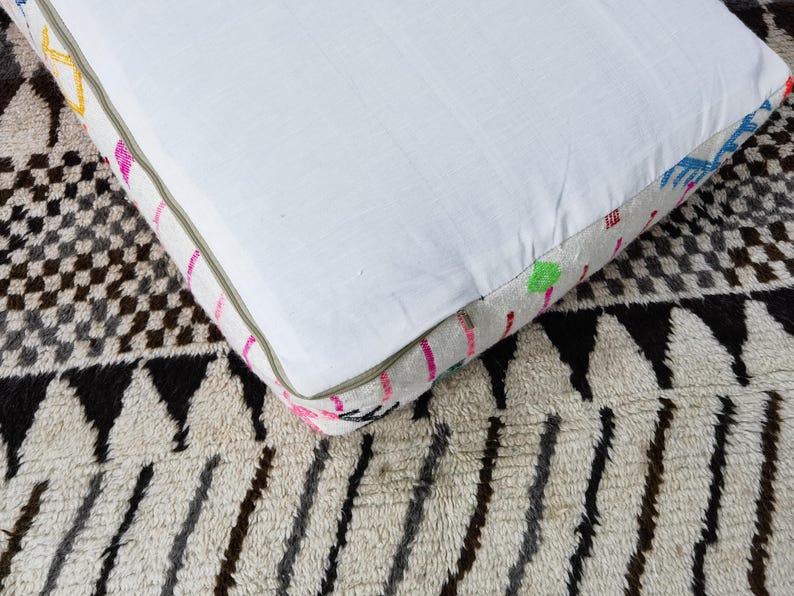 Puede incluir: Un coj&iacute;n cuadrado blanco con un borde estampado colorido. El borde presenta detalles en rosa, amarillo, verde y azul. El coj&iacute;n est&aacute; colocado sobre una alfombra estampada con dise&ntilde;os geom&eacute;tricos en negro, blanco y gris.