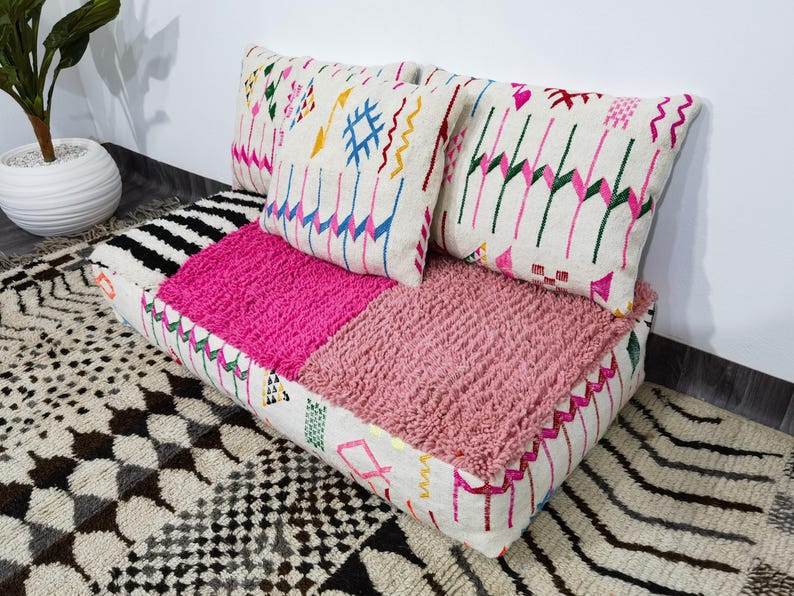 Puede incluir: Un coj&iacute;n decorativo para el suelo con dos cojines a juego. El coj&iacute;n presenta un dise&ntilde;o de patchwork con secciones rosas brillantes y rosas texturizadas. Los cojines y los lados del coj&iacute;n tienen patrones bordados coloridos sobre un fondo blanco.