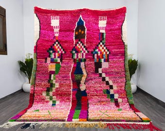 May include: A vibrant, hand-woven rug with a pink and magenta background. The rug features three central, stylized tower-like designs in various colors, including blue, green, and white. The rug has a fringed edge and is displayed on a wooden floor.