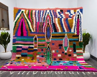 May include: A vibrant, hand-woven rug with a geometric design. The rug features a mix of colors, including pink, blue, green, and yellow. The design includes abstract shapes and patterns. The rug has a fringe along the bottom edge.