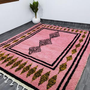 Op de afbeelding: Een roze en zwart Marokkaans tapijt met een geometrisch patroon. Het tapijt heeft een franje rand en is gemaakt van wol.