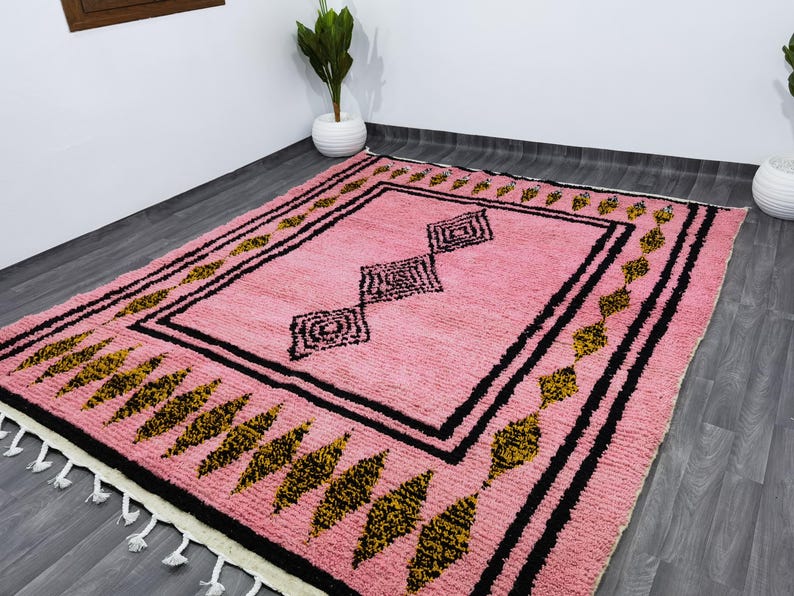 Op de afbeelding: Een roze en zwart Marokkaans tapijt met een geometrisch patroon. Het tapijt heeft een franje rand en is gemaakt van wol.