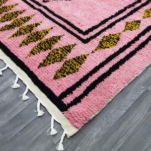Op de afbeelding: Roze en zwart tapijt met een geometrisch patroon en witte franjes. Het tapijt heeft een zwarte rand met een herhalend patroon van gouden en zwarte bladeren.