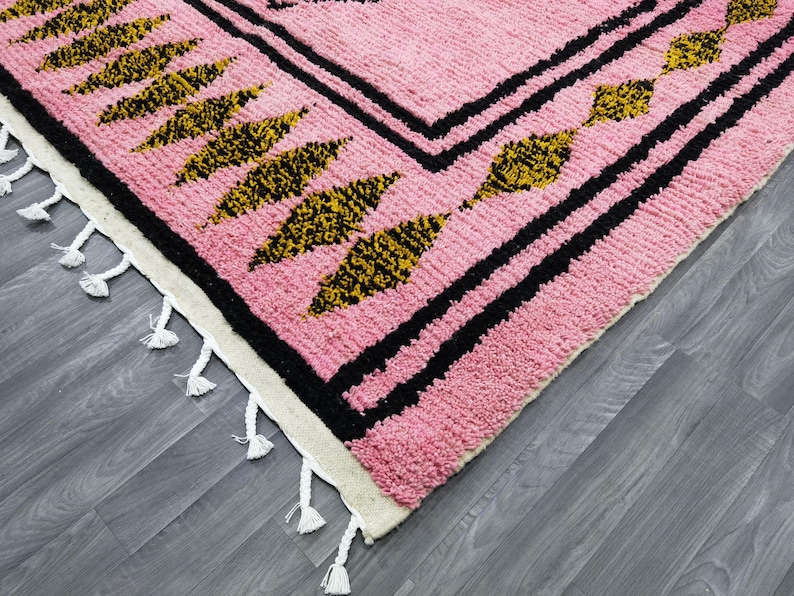 Op de afbeelding: Roze en zwart tapijt met een geometrisch patroon en witte franjes. Het tapijt heeft een zwarte rand met een herhalend patroon van gouden en zwarte bladeren.