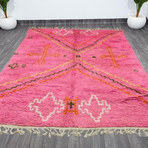 May include: A pink Moroccan rug with a geometric pattern in shades of orange, green, and white. The rug has a fringe border.