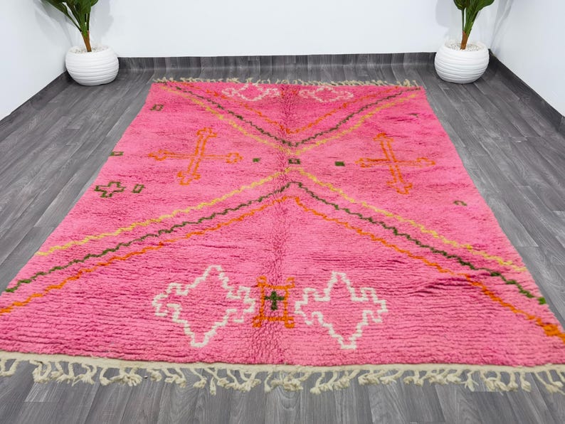 May include: A pink Moroccan rug with a geometric pattern in shades of orange, green, and white. The rug has a fringe border.