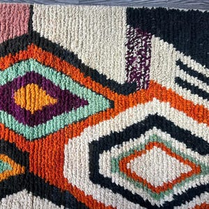 May include: A colorful, abstract rug with a geometric pattern in shades of orange, green, purple, black, and white. The rug has a fringe border.