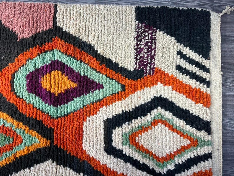 May include: A colorful, abstract rug with a geometric pattern in shades of orange, green, purple, black, and white. The rug has a fringe border.