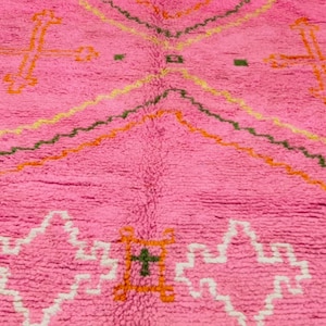 May include: Pink Moroccan rug with a geometric pattern in orange, green, and white. The rug has a fluffy texture and is made of wool.