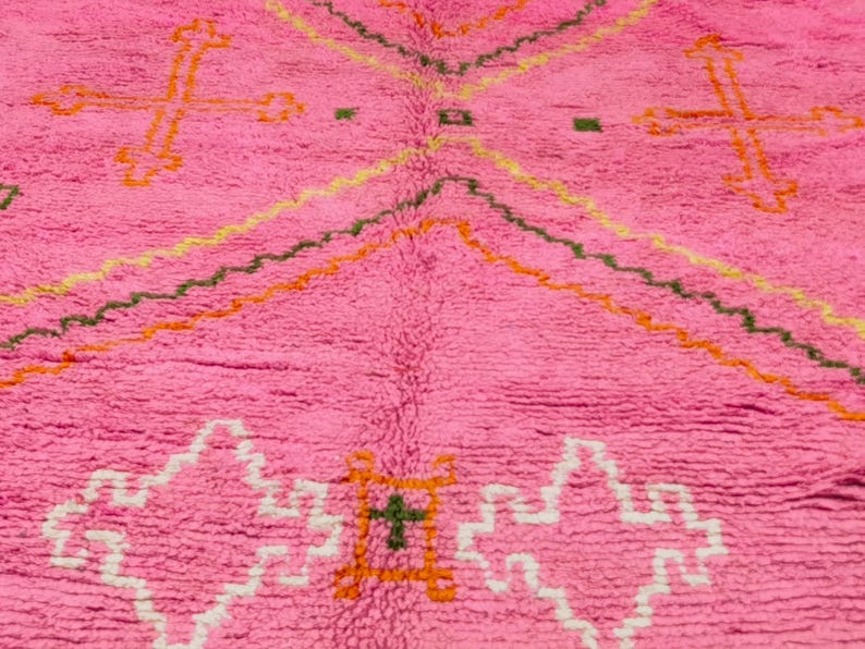 May include: Pink Moroccan rug with a geometric pattern in orange, green, and white. The rug has a fluffy texture and is made of wool.