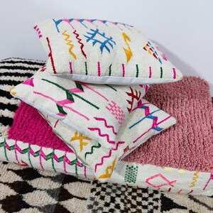 Puede incluir: Una pila de cojines decorativos con diversas texturas y patrones. Los cojines superiores son blancos con dise&ntilde;os bordados coloridos. Tambi&eacute;n se ve un coj&iacute;n rosa texturizado y un coj&iacute;n a rayas blancas y negras.