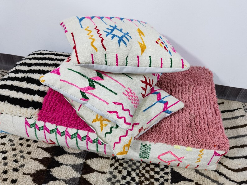 Puede incluir: Una pila de cojines decorativos con diversas texturas y patrones. Los cojines superiores son blancos con dise&ntilde;os bordados coloridos. Tambi&eacute;n se ve un coj&iacute;n rosa texturizado y un coj&iacute;n a rayas blancas y negras.