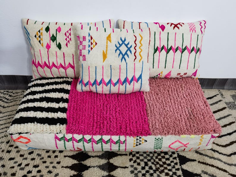 Puede incluir: Un conjunto de cojines decorativos y un coj&iacute;n de suelo. Los cojines tienen un fondo crema con patrones geom&eacute;tricos coloridos en rosa, azul, verde y amarillo. El coj&iacute;n de suelo presenta una secci&oacute;n a rayas blancas y negras, una secci&oacute;n rosa brillante y una secci&oacute;n rosa claro.