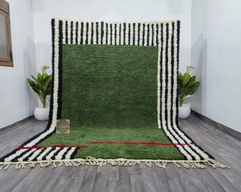 May include: A large, rectangular green rug with a black and white striped border. The rug has a central green field with a red accent line. The rug is made of a textured material and has a fringe along the bottom edge.