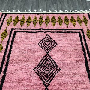 Op de afbeelding: Roze en zwart Marokkaans tapijt met een ruitpatroon. Het tapijt heeft een franje rand.