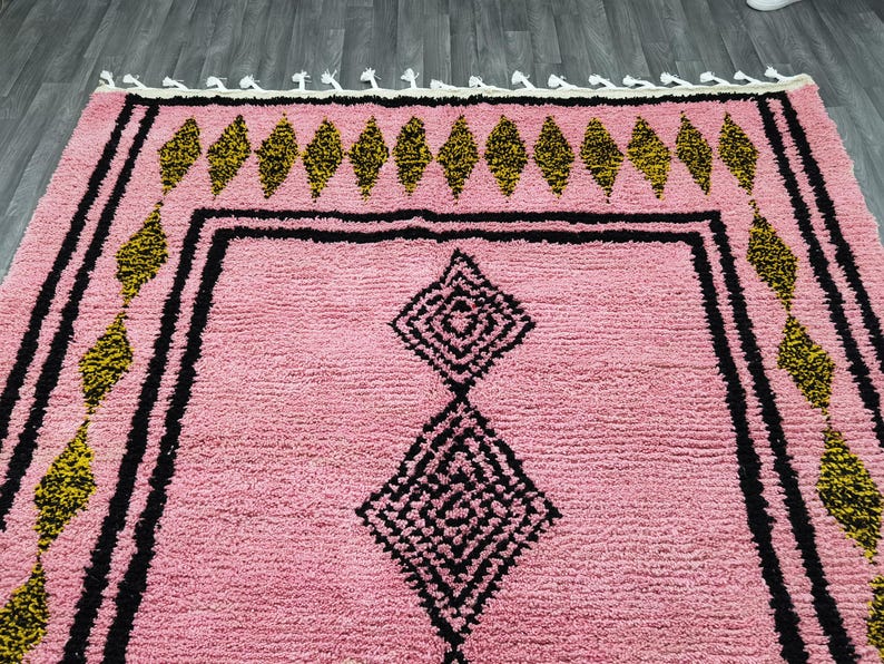 Op de afbeelding: Roze en zwart Marokkaans tapijt met een ruitpatroon. Het tapijt heeft een franje rand.