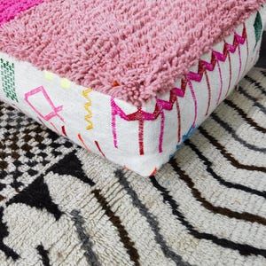 Puede incluir: Un puf marroqu&iacute; rosa y blanco con una parte superior texturizada y bordes bordados. El puf se encuentra sobre una alfombra estampada con rayas negras, grises y marrones y dise&ntilde;os geom&eacute;tricos.