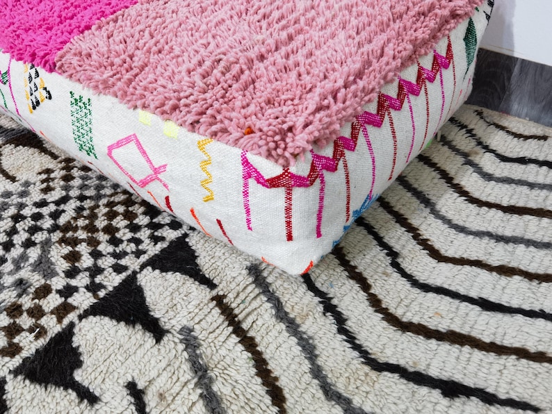Puede incluir: Un puf marroqu&iacute; rosa y blanco con una parte superior texturizada y bordes bordados. El puf se encuentra sobre una alfombra estampada con rayas negras, grises y marrones y dise&ntilde;os geom&eacute;tricos.