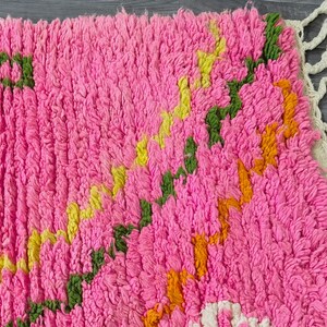 May include: A pink, green, yellow, and orange hand-woven rug with a fringe border. The rug has a geometric pattern.