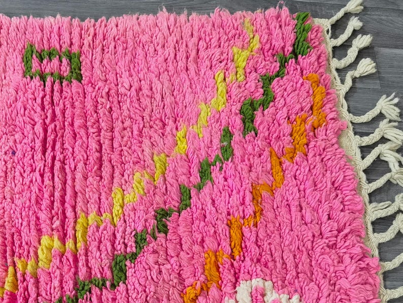 May include: A pink, green, yellow, and orange hand-woven rug with a fringe border. The rug has a geometric pattern.