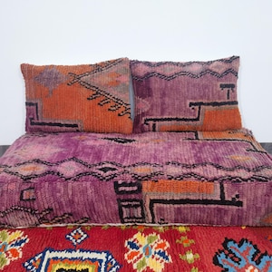 Puede incluir: Un juego de cojines de suelo de estilo marroquí en morado y naranja. El juego incluye un cojín rectangular grande y dos cojines cuadrados más pequeños. Los cojines presentan patrones geométricos en tonos morado, naranja y negro.